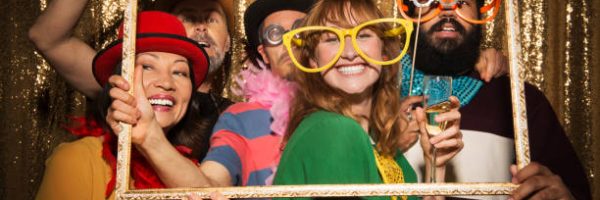 Portrait of cheerful male and female friends wearing party props and taking funny photo with picture frame in photo booth.